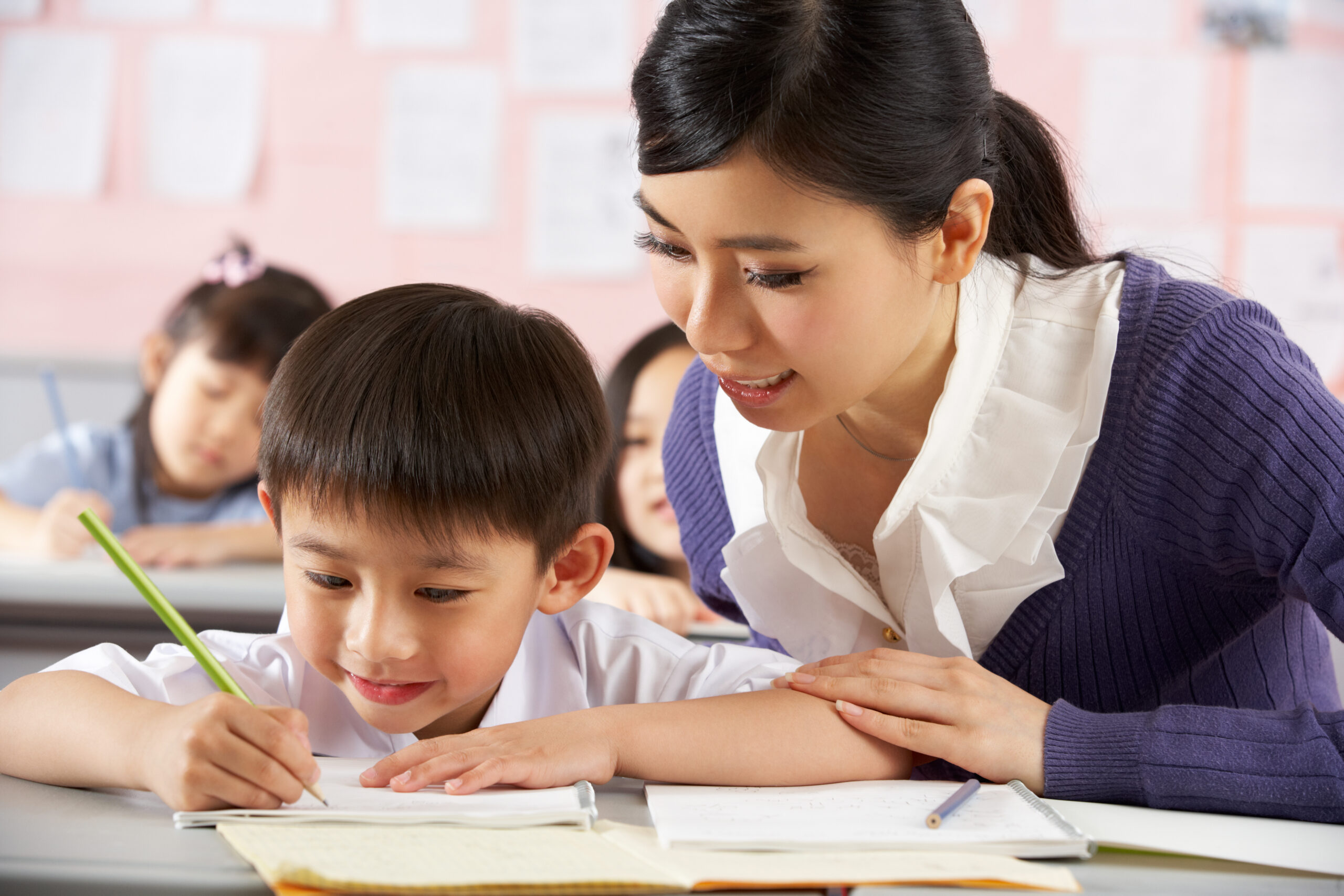 Teacher Helping Student Working At Desk In Chinese School Classroom Sitting Down Checking Work